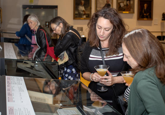 Two Visitors With Drinks Looking At The Healing Words Exhibition